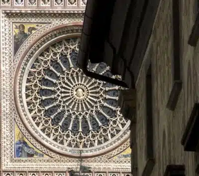 Rose window Duomo di Orvieto – Orvieto | Orvietoviva Ornate Circular Rose Window with Radiating Stone Tracery and Mosaic Surround, Viewed from a Narrow Street with a Gutter in the Foreground.