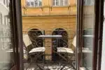 terrazzino-palazzaccio – Orvieto | Orvietoviva Open French Doors Reveal a Small Wrought-iron Balcony with Two Beige Folding Chairs and a Metal Bistro Table, Overlooking a Yellow Building.