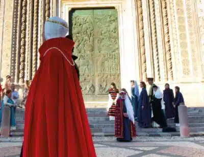 corteo-delle-dame-orvieto – Orvieto | Orvietoviva a Group of People in Medieval Robes Outside an Ornate Building Doorway; a Person in a Bright Red Cloak Dominates the Foreground.
