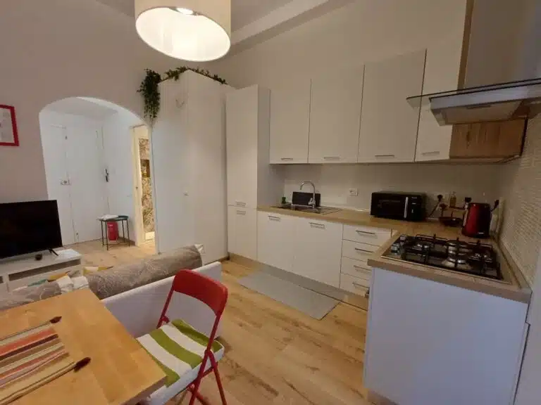 Open-plan kitchen with white cabinets, wooden countertops, gas stove, and a small dining table with a red chair in a cozy living area.