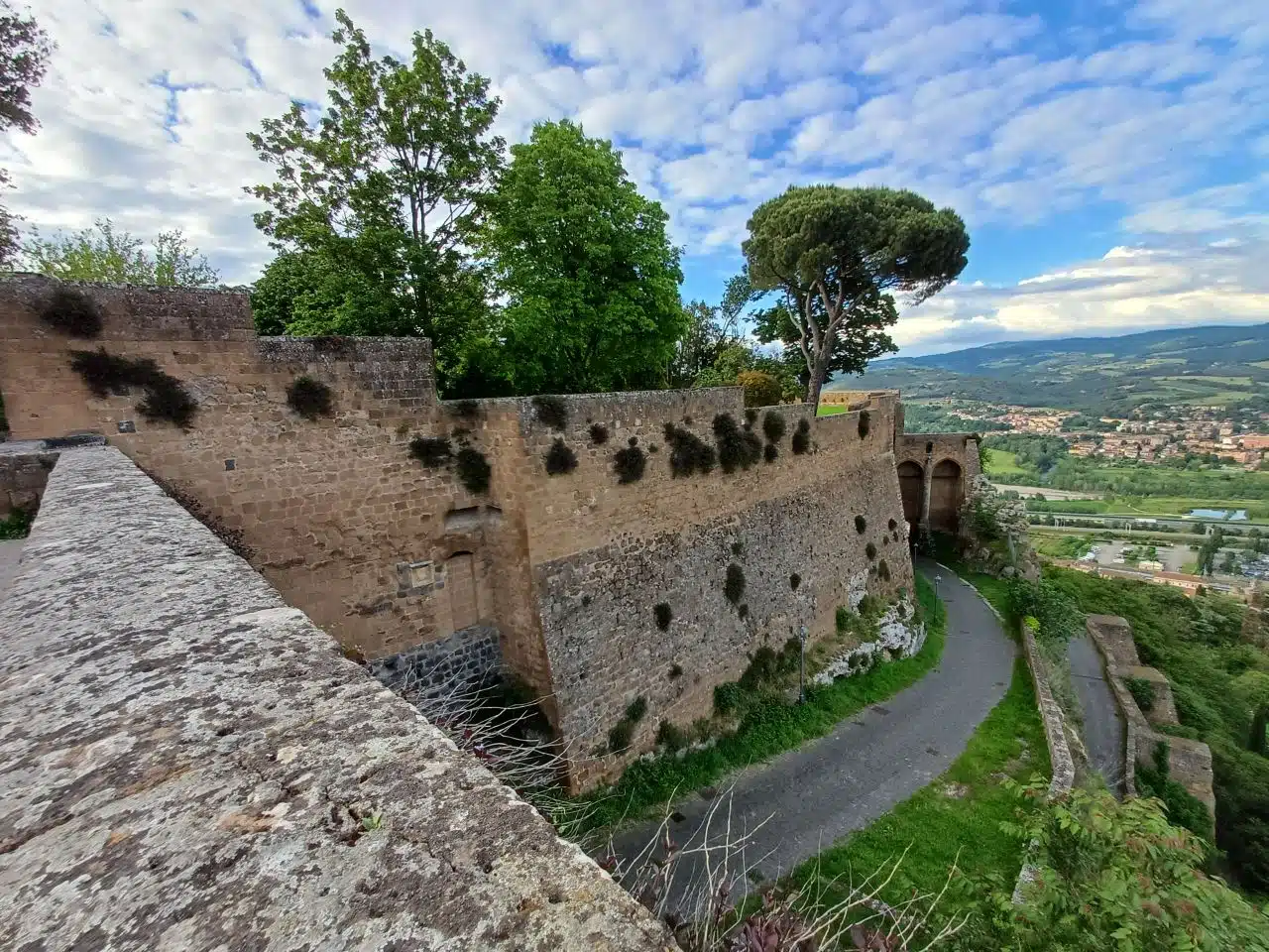 piagge-orvieto – Orvieto | Orvietoviva Historic Stone Fortification Along a Winding Hillside Path, with Trees and a Valley Town in the Distance Under a Partly Cloudy Sky. Orvietoviva - Informazioni Turistiche, Eventi Della Città Umbra Di Orvieto. Consigli Per Visitare Orvieto, Cosa Vedere E Fare a Orvieto, Dormire E Mangiare
